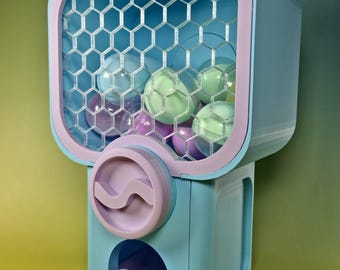 May include: A light blue and lavender gumball machine featuring a clear hexagonal grid front panel. The machine is filled with several translucent spherical capsules in pastel shades of green, blue, and purple, with one capsule resting in the tray.