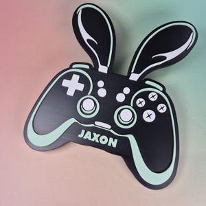 May include: Black and white controller with bunny ears, featuring a plus sign, circles, and crosses. The name "JAXON" is displayed in white. The controller has a mint green outline and accents.