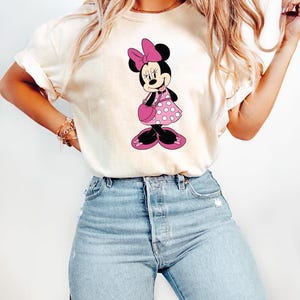 Puede incluir: Camiseta color crema con un gr&aacute;fico de Minnie Mouse. Minnie est&aacute; representada con un vestido rosa de lunares, un lazo a juego y un bolso. La camiseta se combina con unos vaqueros azules de lavado claro, creando un look casual y cotidiano.