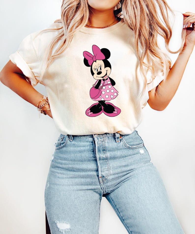 Puede incluir: Camiseta color crema con un gr&aacute;fico de Minnie Mouse. Minnie est&aacute; representada con un vestido rosa de lunares, un lazo a juego y un bolso. La camiseta se combina con unos vaqueros azules de lavado claro, creando un look casual y cotidiano.