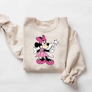 Puede incluir: Sudadera beige con un gr&aacute;fico de Minnie Mouse en rosa, negro y blanco. Minnie Mouse se representa sosteniendo una varita en forma de estrella y usando un lazo rosa, un vestido y zapatos. La sudadera tiene mangas largas.