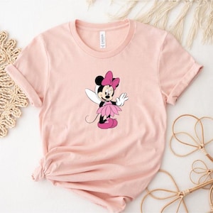 Puede incluir: Camiseta rosa claro con un dise&ntilde;o de Minnie Mouse de dibujos animados como hada. Minnie Mouse lleva un lazo rosa, un tut&uacute; rosa y zapatos rosas. La camiseta tiene las mangas dobladas.