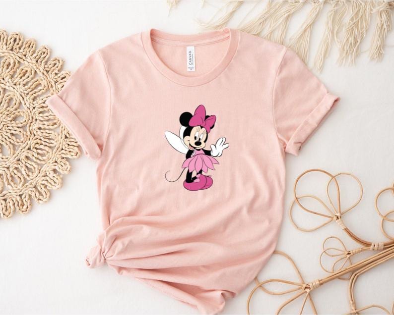 Puede incluir: Camiseta rosa claro con un dise&ntilde;o de Minnie Mouse de dibujos animados como hada. Minnie Mouse lleva un lazo rosa, un tut&uacute; rosa y zapatos rosas. La camiseta tiene las mangas dobladas.