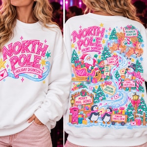 May include: A white crewneck sweatshirt featuring a colorful graphic print. The front displays the text "NORTH POLE HOLIDAY DISPATCH" in pink. The back shows a detailed holiday scene with Santa, reindeer, a snowman, and penguins with text.