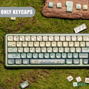 May include: A custom keyboard with light green and off-white keycaps. The keycaps feature illustrations of trees, frogs, and other nature-themed designs. The keyboard is surrounded by moss and other keycaps.