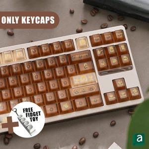 Puede incluir: Un teclado con teclas marrones y beige, algunas con iconos temáticos de café. Las teclas tienen un aspecto translúcido. La imagen incluye el texto "ONLY KEYCAPS" y "FREE FIDGET TOY".