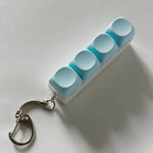 May include: A light blue keyboard keycap keyring with four keycaps. The keycaps are attached to a white base and connected to a silver keyring with a lobster clasp.