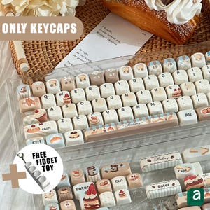 Puede incluir: Un juego de teclas de teclado con temática de panadería. Las teclas son blancas con varios diseños de postres, incluyendo pasteles, bollería y pan. La palabra "Bakery" está impresa en la barra espaciadora. La imagen también incluye un juguete antiestrés gratis.