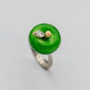 May include: A ring featuring a glossy, bright green apple-shaped centerpiece. A small, marquise-cut clear gemstone is set in gold at the top of the apple, next to a small gold stem. The ring band is made of a smooth, silver-toned metal material.