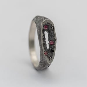 Puede incluir: Un anillo de plata oxidada oscura con una superficie texturizada y rugosa. El centro alargado y hueco presenta un grupo de pequeños rubíes redondos de color rojo intenso y diminutas piedras negras incrustadas en el acabado metálico oscuro.