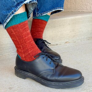 May include: A pair of black leather lace-up shoes with a smooth finish and thick soles, worn with mid-calf knit socks featuring a rust-orange body, a teal cuff, and a ribbed texture, paired with blue denim jeans with frayed hems at the bottom.