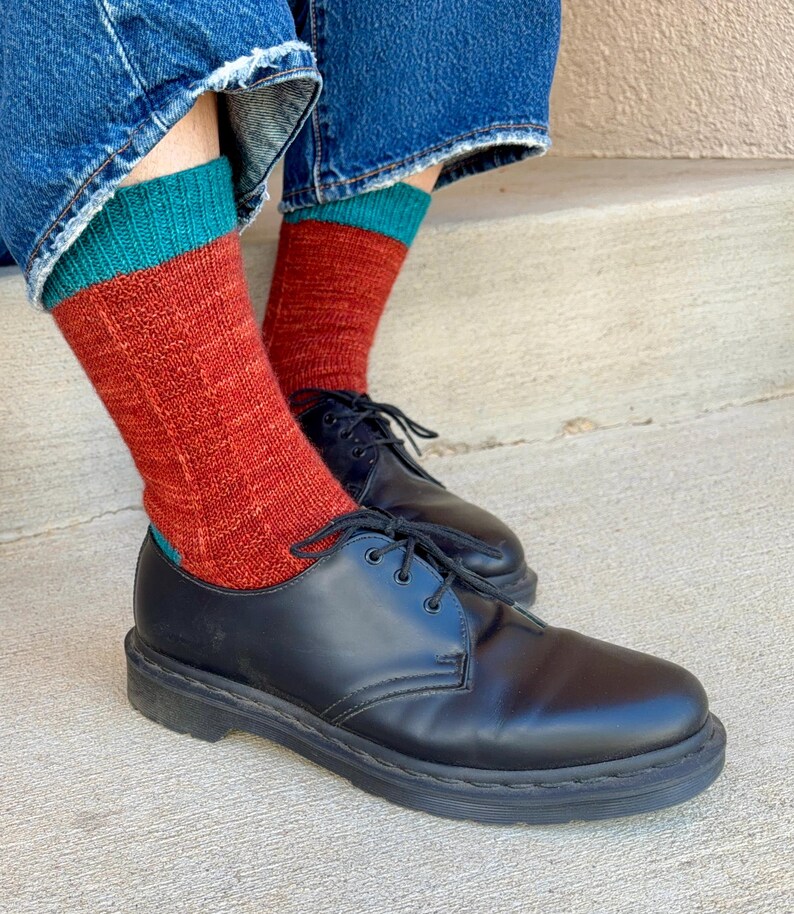 May include: A pair of black leather lace-up shoes with a smooth finish and thick soles, worn with mid-calf knit socks featuring a rust-orange body, a teal cuff, and a ribbed texture, paired with blue denim jeans with frayed hems at the bottom.