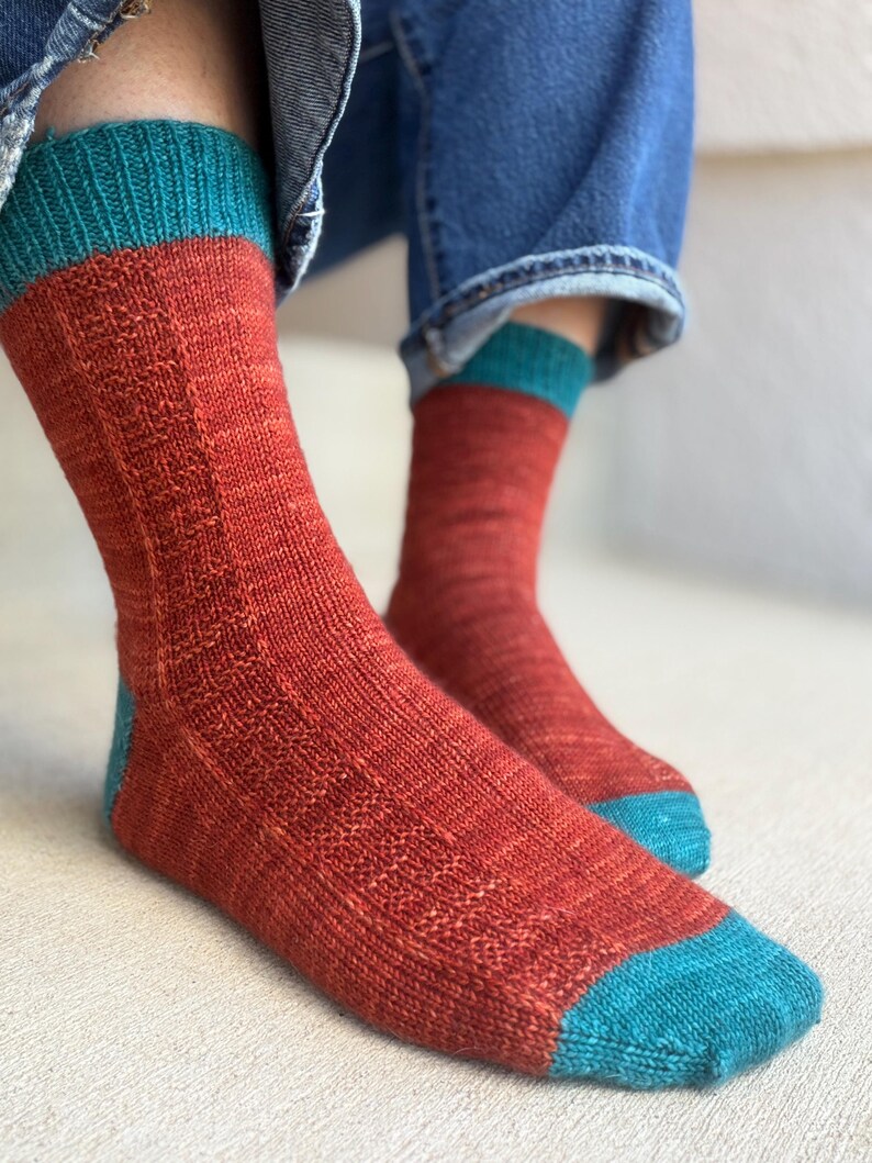May include: A pair of hand-knitted socks featuring a vibrant rust-orange main body with a textured vertical stitch pattern. The socks have contrasting teal cuffs, heels, and toe sections, creating a distinct color-blocked appearance.
