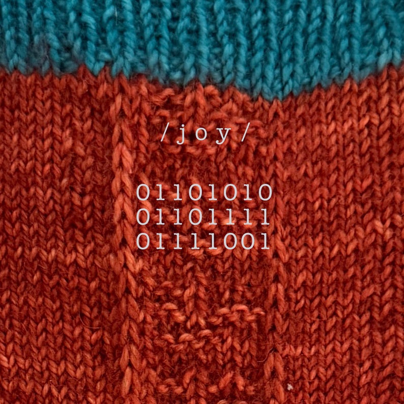 May include: A knitted fabric featuring a horizontal band of teal yarn at the top and a textured rust-orange section below. The rust-orange area displays the text /joy/ followed by three lines of binary code: 01101010, 01101111, and 01111001.