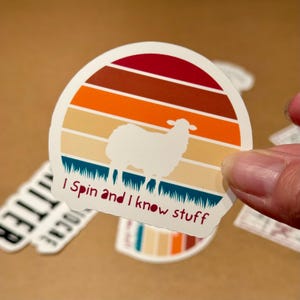 May include: A white sheep silhouette sticker with the text "I Spin and I know stuff." The sticker has a retro sunset design with red, orange, and tan stripes. The bottom of the sticker has a blue grass design.