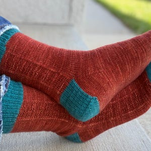 May include: A pair of knitted socks featuring a rust-colored main body with teal accents on the cuffs, heels, and toes. The socks have a ribbed texture along the leg and a smooth knit finish on the foot, worn with blue denim jeans.