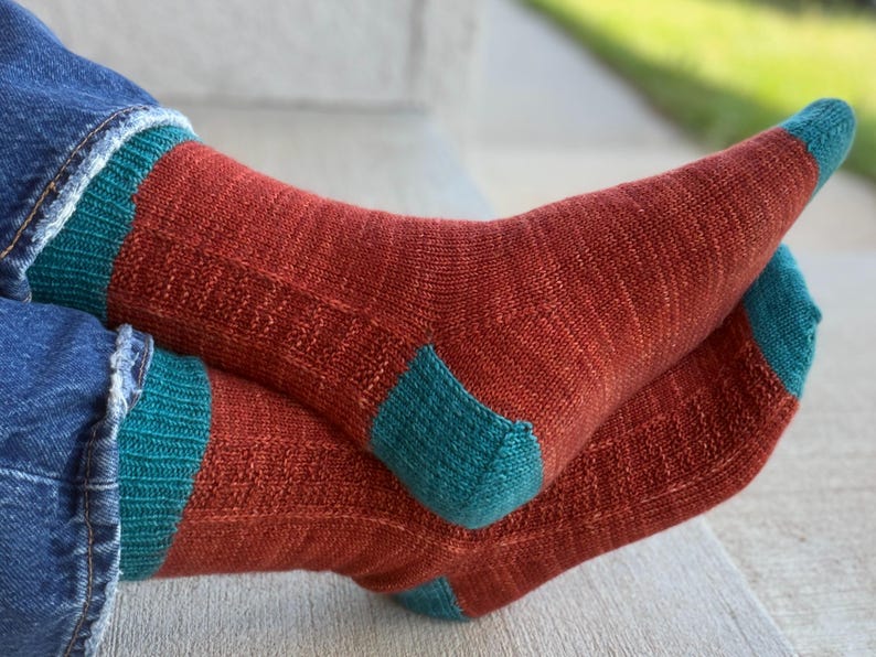 May include: A pair of knitted socks featuring a rust-colored main body with teal accents on the cuffs, heels, and toes. The socks have a ribbed texture along the leg and a smooth knit finish on the foot, worn with blue denim jeans.
