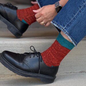 May include: A pair of black leather lace-up shoes paired with mid-calf knit socks. The socks feature a textured rust-colored body with a solid teal cuff at the top. The shoes have a smooth finish, rounded toes, and black laces, worn with blue denim jeans.