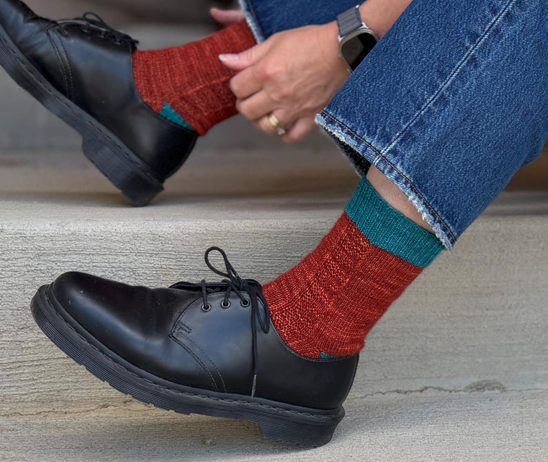 May include: A pair of black leather lace-up shoes paired with mid-calf knit socks. The socks feature a textured rust-colored body with a solid teal cuff at the top. The shoes have a smooth finish, rounded toes, and black laces, worn with blue denim jeans.