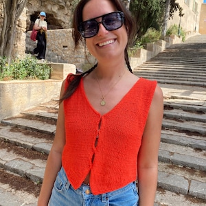 Könnte beinhalten: Eine leuchtend orangefarbene Strickweste mit V-Ausschnitt und Knopfverschluss. Die Weste wird mit hellblauen Jeans kombiniert. Die Person trägt eine schwarze Sonnenbrille und eine goldene Halskette. Der Hintergrund zeigt Steinstufen und Grün.