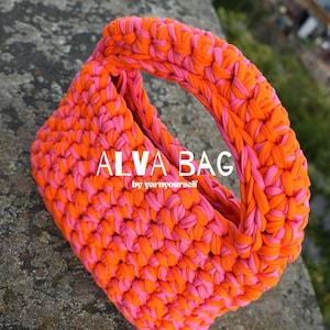 Op de afbeelding: Een gehaakte handtas gemaakt van dik, verweven feloranje en felroze garen. De tas heeft een ronde vorm met een geïntegreerd handvat en toont de witte tekst ALVA BAG by yarnyourself gecentreerd op de voorzijde van het oppervlak.