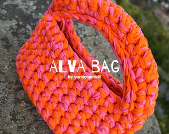 May include: A crochet handbag crafted from thick, intertwined bright orange and hot pink yarn. The bag features a rounded shape with an integrated handle and displays the white text ALVA BAG by yarnyourself centered on the front surface.