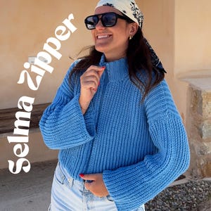 Könnte beinhalten: Ein leuchtend blauer Rippstrickpullover mit hohem Kragen und Reißverschluss auf der Vorderseite. Der Text 'Selma Zipper' ist in weißer Schrift auf der linken Seite zu sehen. Der Pullover hat lange, weit geschnittene Ärmel und ein Strickmuster.