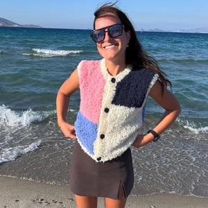 Könnte beinhalten: Eine ärmellose, geknöpfte Weste aus texturiertem, flauschigem Material mit einem Color-Block-Muster in Pink, Marineblau, Hellblau und Creme. Die Weste hat einen Rundhalsausschnitt und vier dunkle Knöpfe entlang der vorderen Verschlussleiste.