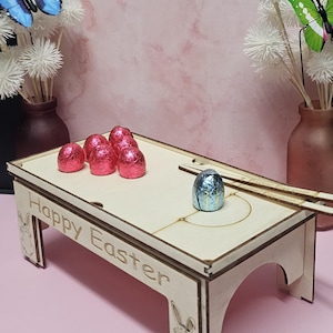 May include: Wooden Easter game with a blue and gold foil-wrapped chocolate egg in the center of the game board. The game board has two wooden sticks and the words "Happy Easter" engraved on the side. There are six pink foil-wrapped chocolate eggs on the game board.