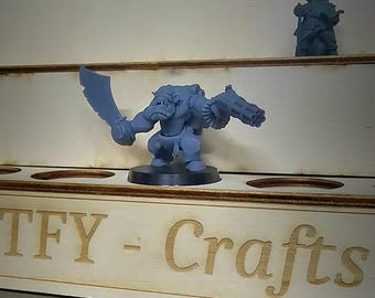 May include: A grey plastic miniature of an Orc warrior holding a sword and a gun. The miniature is on a black base and is displayed on a wooden shelf with the text "TFY - Crafts" engraved on the front.