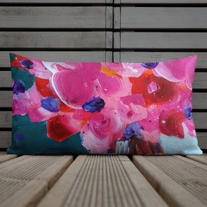 May include: Rectangular decorative pillow with a vibrant abstract floral design. The pillow features large brushstrokes in shades of pink, red, and purple against a teal and light blue background. The pillow is placed on a wooden surface.
