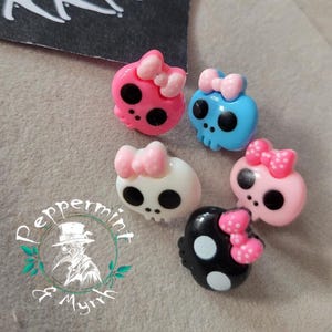 May include: A collection of colorful skull-shaped charms with bows. The charms are in various colors, including pink, blue, white, and black. Each skull has large black eye sockets and a bow on top. The charms are likely for jewelry or craft projects.