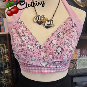 May include: Pink halter top with a Hello Kitty design. The top has a pink and white gingham trim and a pink halter strap. A necklace with the words "YOU'RE SO COOL" is visible. The top is from Dirty Martini Clothing.