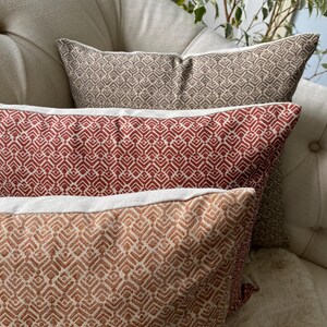May include: Close-up of three decorative throw pillows with geometric patterns. The pillows are in shades of red, brown, and tan, with white trim. They are arranged on a beige upholstered chair, creating a cozy and stylish home decor scene.