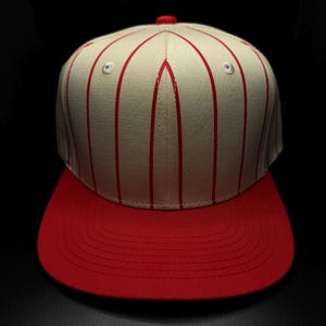 Puede incluir: Gorra de béisbol con corona crema con rayas verticales rojas y visera roja. La gorra tiene una parte superior redondeada con un pequeño botón rojo y dos ojales a cada lado. La visera es plana y de color rojo sólido.