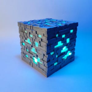 Peut inclure: Une lampe en forme de cube gris avec un design pixelisé et des lumières turquoise brillantes à l'intérieur. La lampe est en plastique et a une surface texturée.