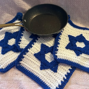 May include: A black frying pan rests on three white and blue crochet potholders. Each potholder features a blue Star of David design. The potholders have a blue border and are square-shaped.