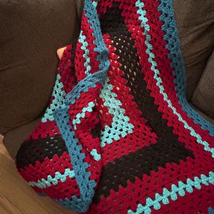 May include: A handmade crochet blanket in a granny square pattern. The blanket features a color scheme of red, black, and turquoise blue. The blanket is folded over a dark gray surface.