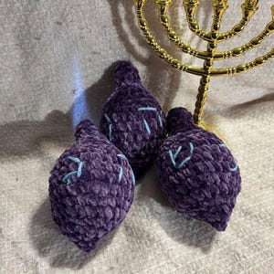 May include: Three plush, purple ornaments with light blue stitching, arranged near a gold menorah. The ornaments have a bulbous shape, and the menorah has nine candle holders. The background is a textured, off-white fabric.