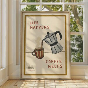 Puede incluir: Una lámina enmarcada con una ilustración dibujada a mano de una cafetera italiana plateada vertiendo café en una taza de rayas rojas y blancas. El texto dice LIFE HAPPENS COFFEE HELPS y SLOW DOWN BREW DEEP STAY KIND en letras rojas oscuras.