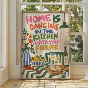May include: A framed wall art print featuring the text "HOME IS DANCING IN THE KITCHEN WITH YOU FOREVER" in colorful hand-lettered typography. The illustration depicts a blue and white striped pitcher with flowers, a bowl of lemons, wine glasses, a bottle of wine, and bread on a green and white striped tablecloth.