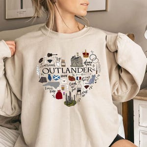 Può includere: Felpa beige con un design a forma di cuore con la scritta "OUTLANDER" al centro. Il cuore è pieno di illustrazioni e testo, tra cui "Sassenach", "dinna fash" e "Frasers Ridge".