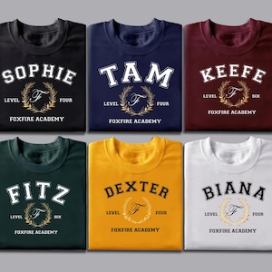 May include: Six folded t-shirts in various colors, each with a name, level, and "Foxfire Academy" text. The names are "Sophie", "Tam", "Keefe", "Fitz", "Dexter", and "Biana". Each shirt has a gold wreath design.