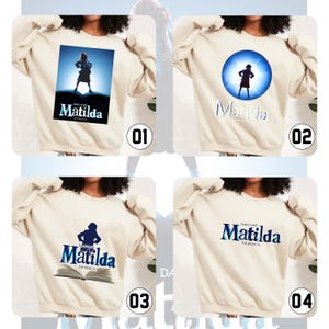 May include: Cream-coloured sweatshirts with designs from the musical "Matilda". The designs include a girl on a stage, a silhouette in a blue circle, and the musical's logo in blue and white.