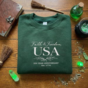 May include: A dark green sweatshirt with the text "Faith & Freedom USA" and "250 Year Anniversary Est. 1776" in white script. The sweatshirt is surrounded by a book, a witch's broom, and potion bottles.