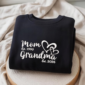 Puede incluir: Sudadera negra con texto blanco "Mom Est. 1990 Grandma Est. 2026" y dos corazones entrelazados. La sudadera está doblada sobre una superficie de color claro.