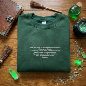 May include: A dark green sweatshirt with white text that reads, "Will you come to my cottage this summer?" and other text. The sweatshirt is on a wooden surface with a book, brooms, and potion bottles.