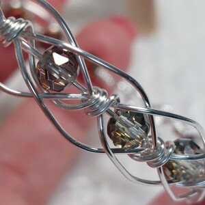 May include: Close-up of a silver wire bracelet with faceted, smoky-coloured beads. The wire is intricately woven, creating a delicate, open design. The beads are evenly spaced, adding a touch of sparkle to the handcrafted jewellery.