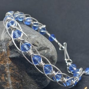 May include: A wire-wrapped bracelet featuring a series of faceted, translucent blue crystal beads secured within a silver-toned metal wire frame. The design consists of interlocking wire loops that hold each bead in place, finished with a hook clasp.