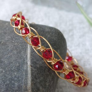 May include: A gold-toned wire-wrapped bracelet featuring a series of faceted, deep red circular beads. The metal wire is intricately coiled around each bead, creating a continuous, repeating pattern that forms the structure of the circular bangle.